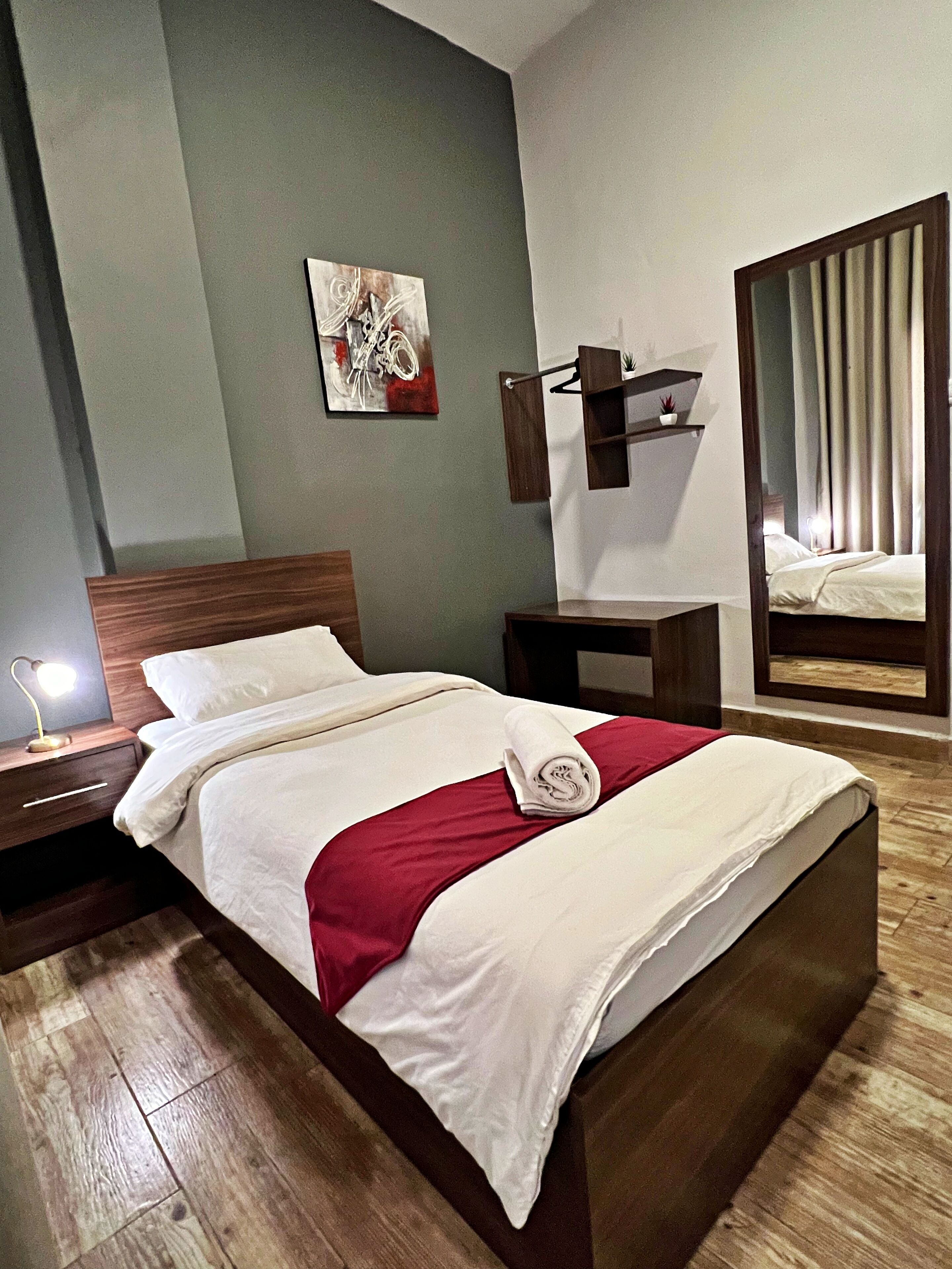 A neatly made bed with white linens and a red accent, a bedside lamp, a wooden nightstand, a wall-mounted shelf with a plant, and a large mirror reflecting a window with curtains.