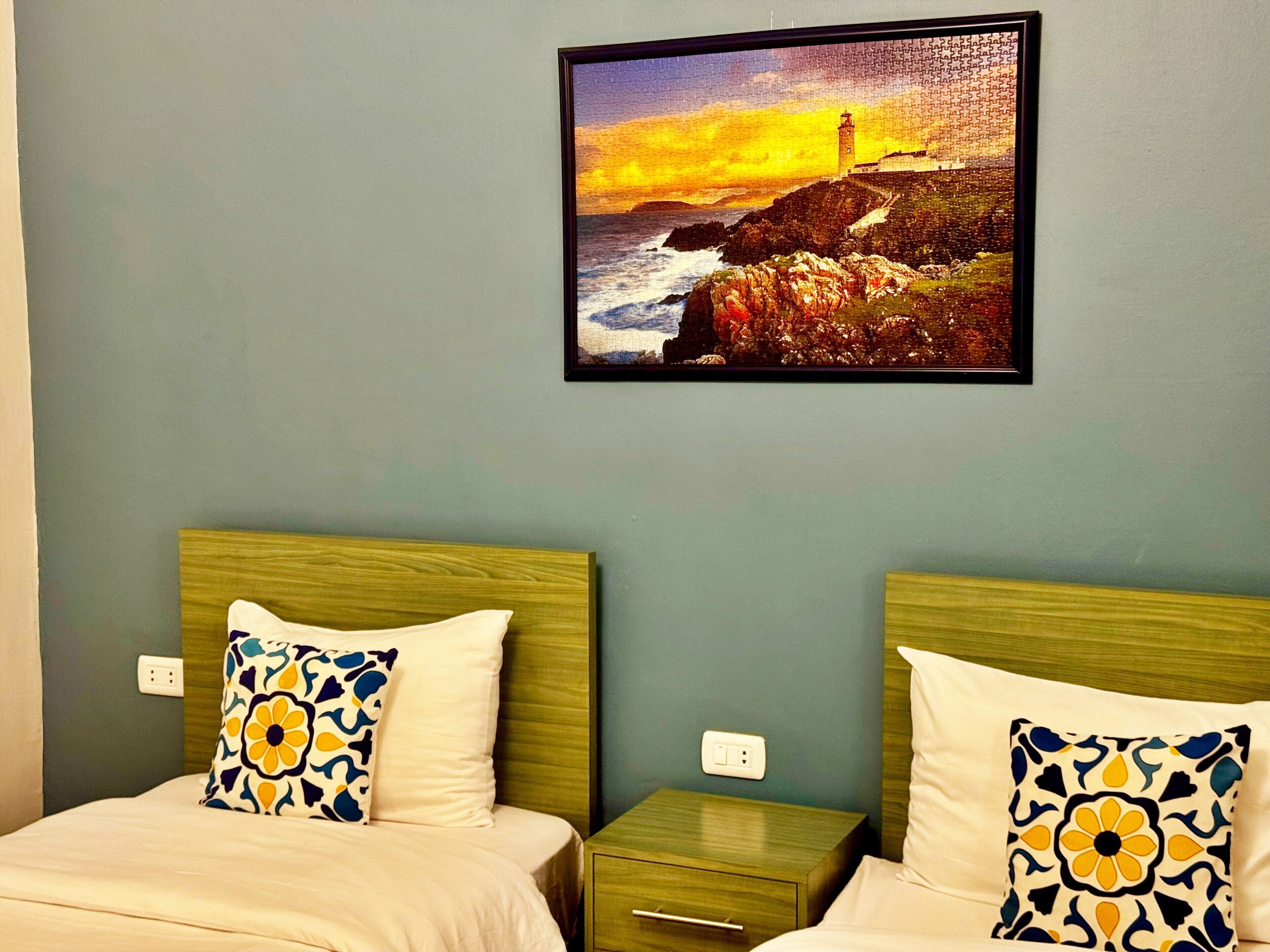 A hotel room with two beds, a nightstand, and a wall-mounted picture of a lighthouse at sunset.