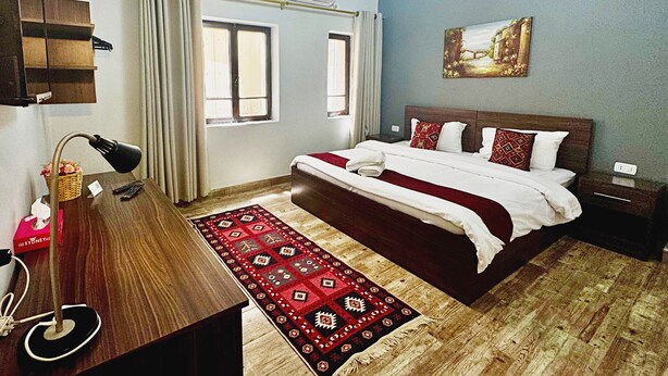 Una habitación de hotel con piso de madera, una cama grande con ropa de cama blanca y roja, un tapete con diseño rojo y negro, un escritorio con lámpara y un cuadro en la pared.