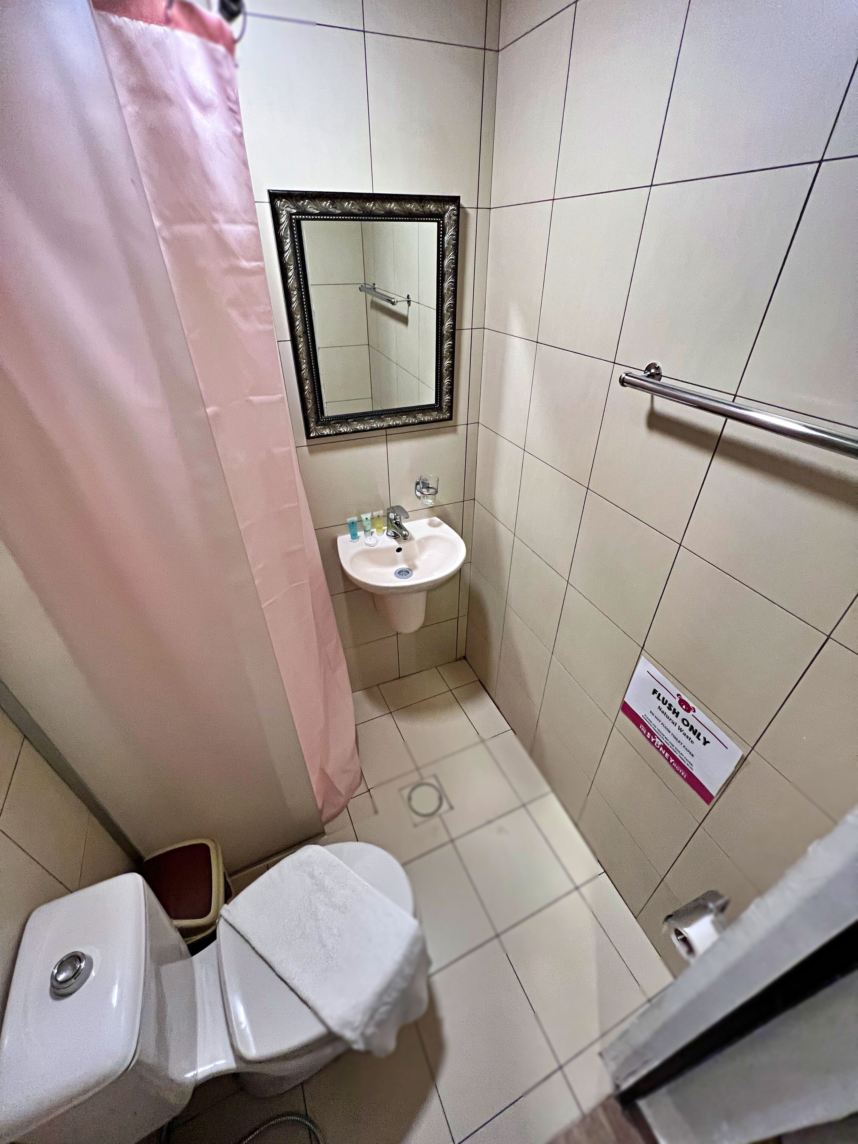 Un baño pequeño con inodoro, lavamanos y espejo. Hay una cortina de ducha rosa y un letrero que indica que solo se puede usar la descarga.