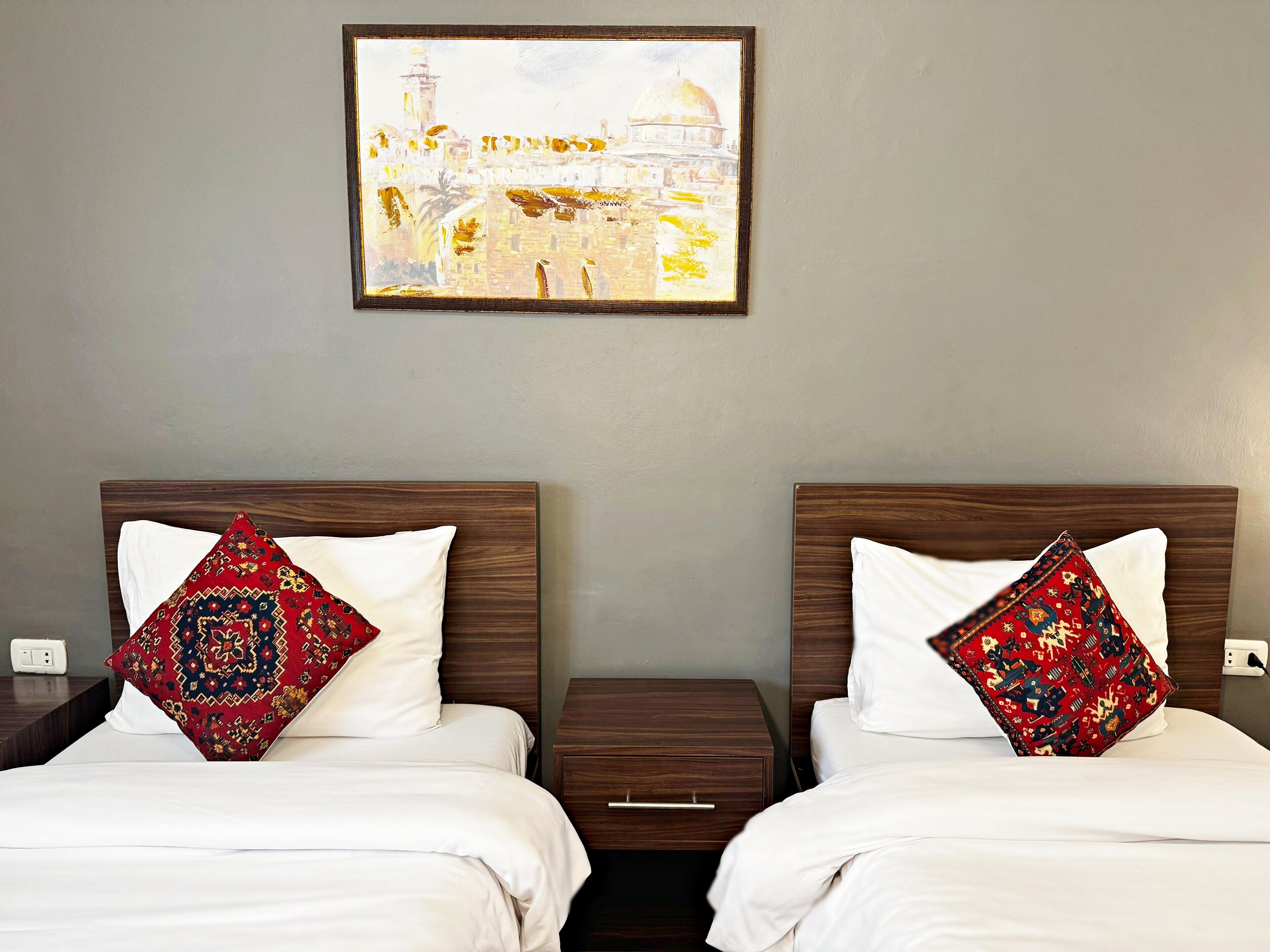 A hotel room with two beds, each with a patterned pillow and a framed cityscape painting above.