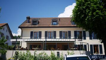 Un bâtiment sur lequel on peut lire l’inscription « Auberge Communale », et qui possède un balcon où se trouvent des personnes.