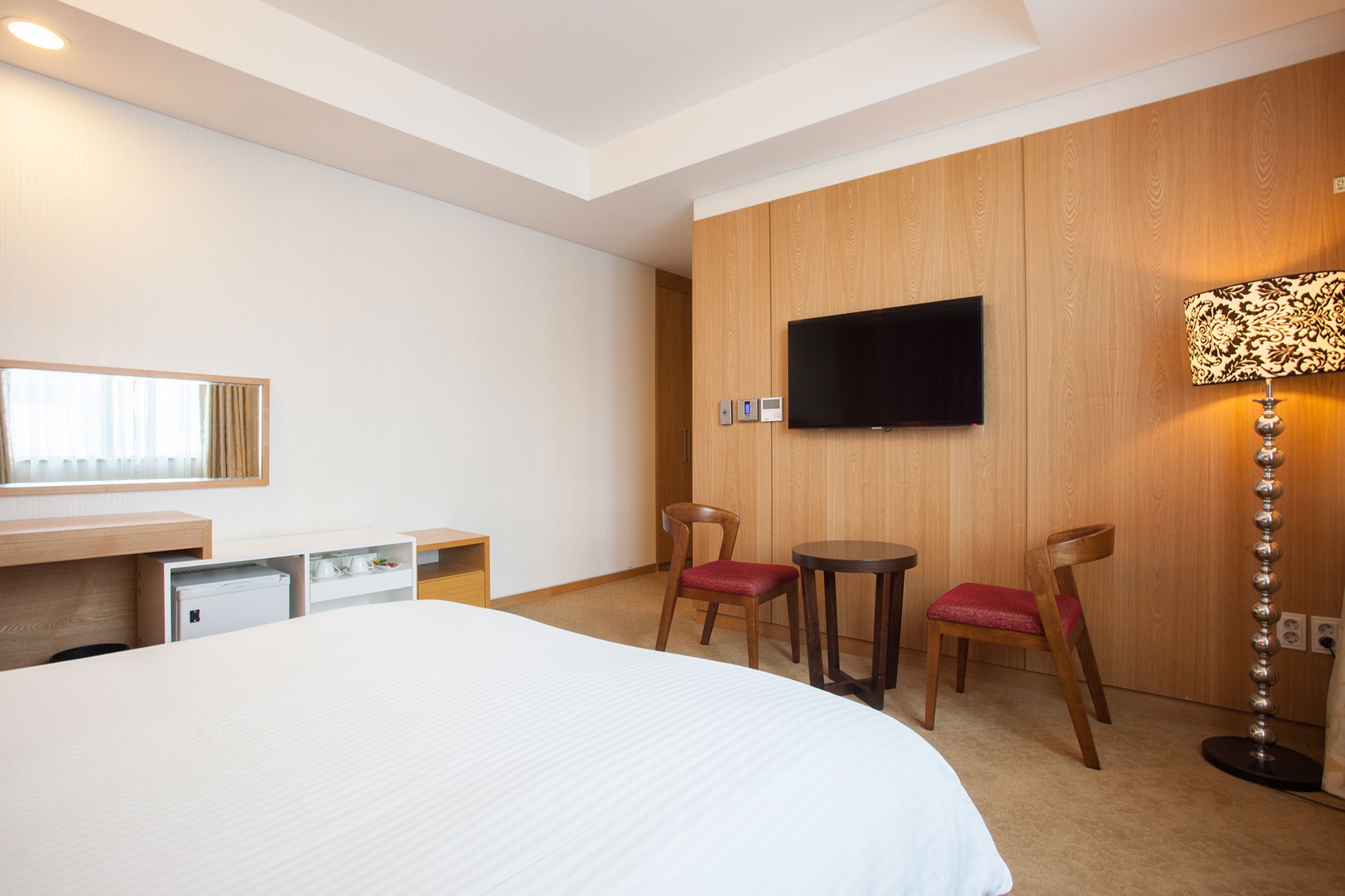 A hotel room with a bed, a desk with a computer, two chairs, a TV mounted on the wall, and a floor lamp.
