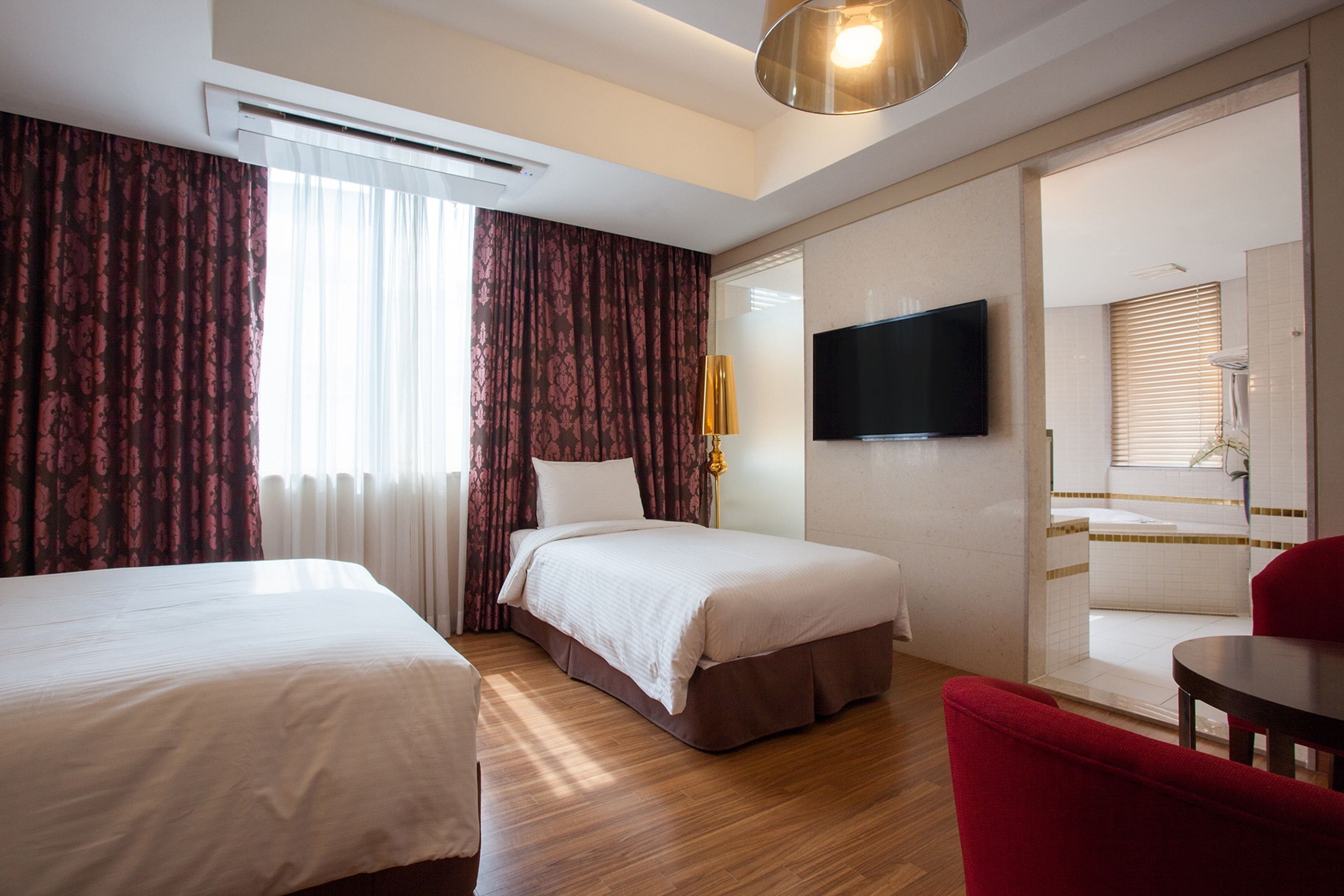 Habitación de hotel con dos camas, un televisor, una silla roja y un baño visible a través de una puerta de cristal.