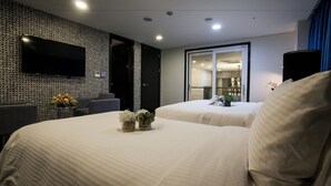 Une chambre d’hôtel avec deux lits, un téléviseur à écran plat, une petite table agrémentée de fleurs et une vue sur la cuisine à travers une porte vitrée.