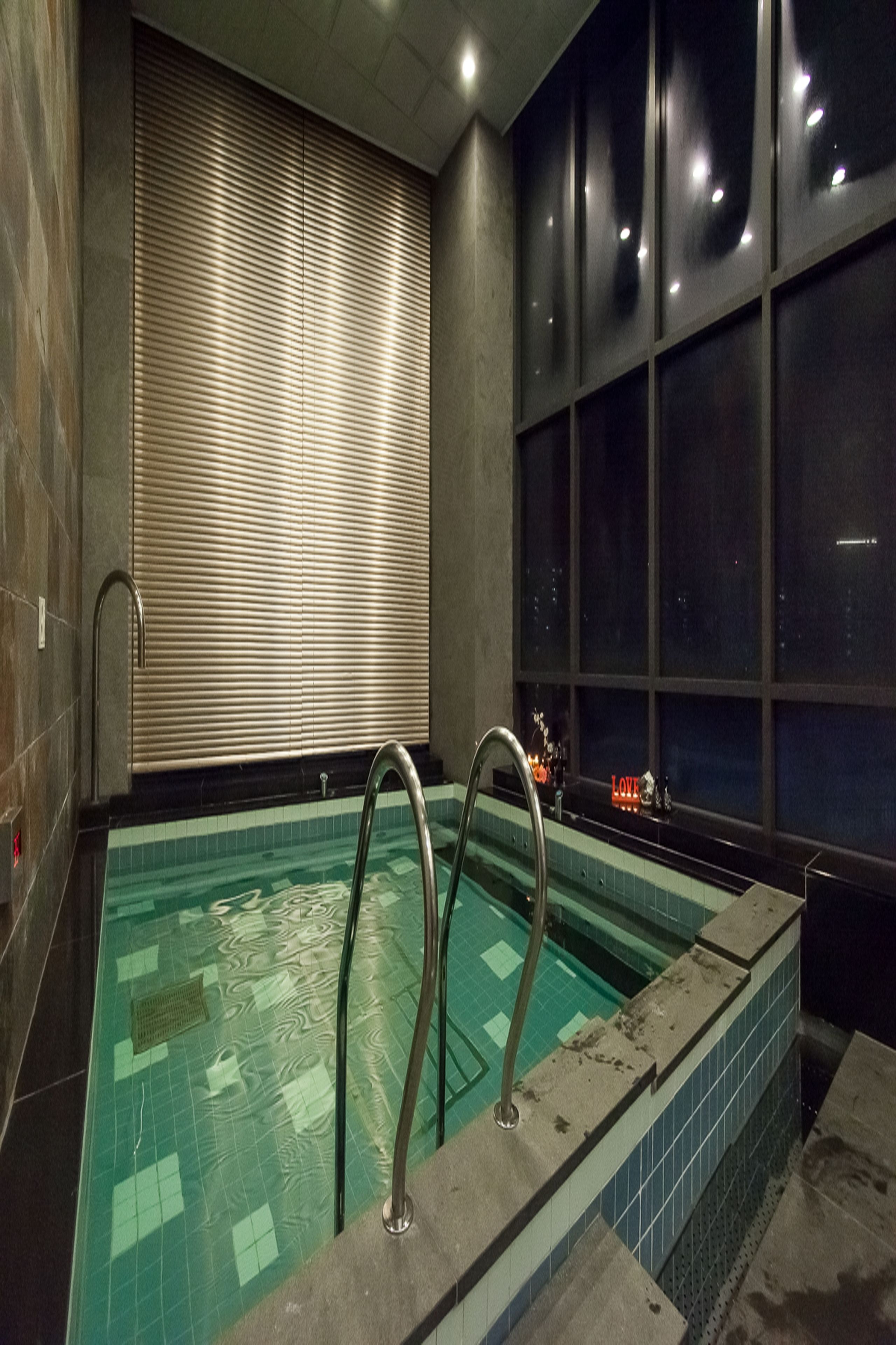A swimming pool with a tiled floor, metal handrails, and a window with blinds.