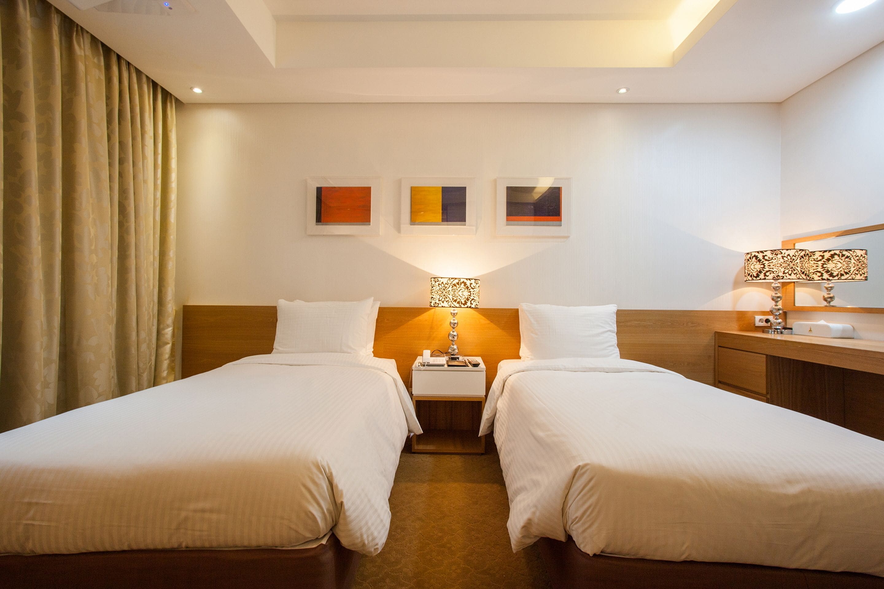 A hotel room with two single beds, a wooden headboard, a nightstand with a lamp, and three framed artworks on the wall.