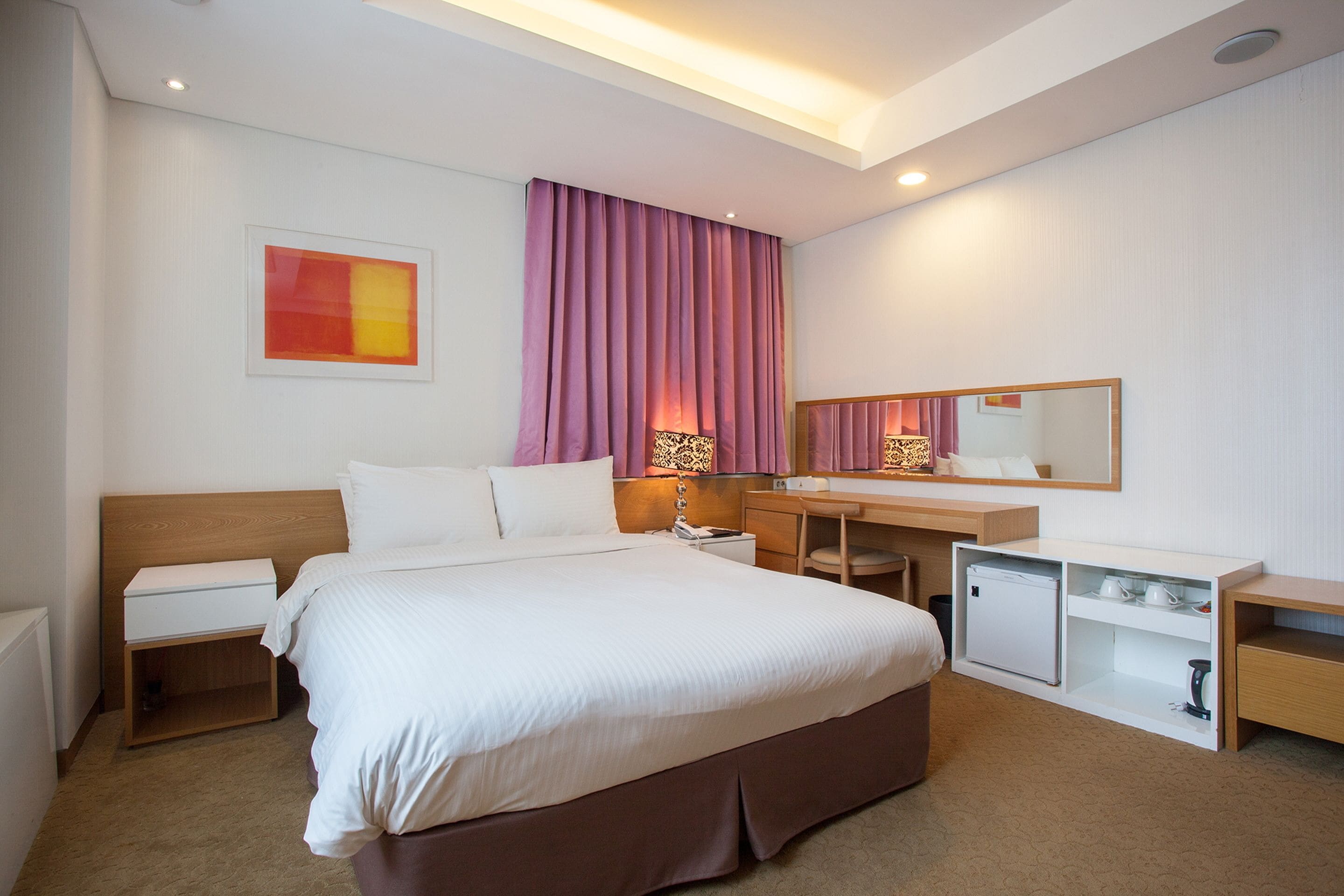 A hotel room with a bed, bedside table, desk, and a small refrigerator.