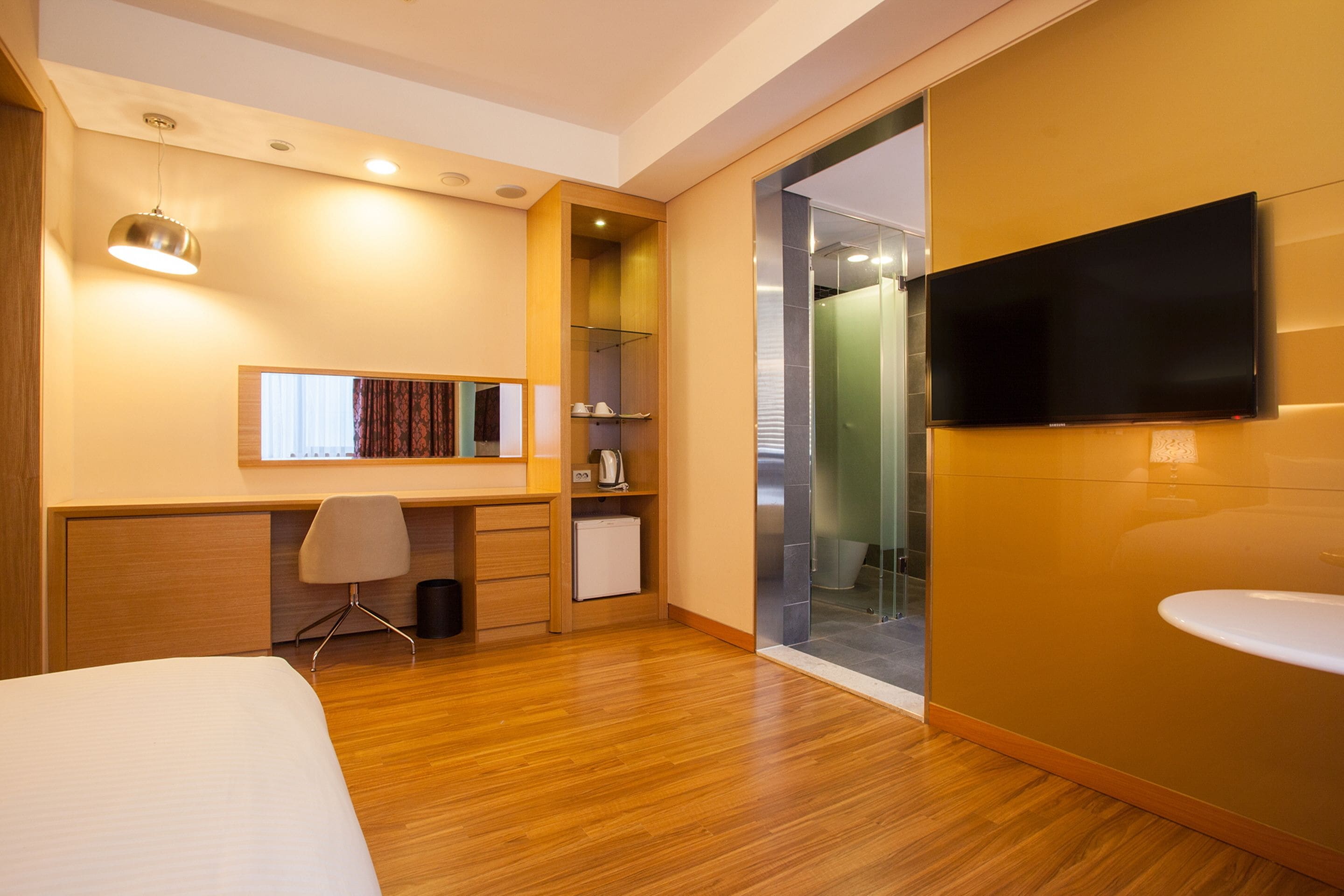 A modern hotel room with a wooden floor, a bed, a desk with a chair, a television mounted on the wall, and a glass shower enclosure.