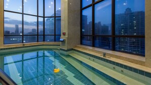 Une grande piscine intérieure aux carreaux bleu clair, avec un canard en caoutchouc jaune flottant à la surface. De grandes fenêtres l'entourent et offrent une vue sur le paysage urbain au crépuscule.