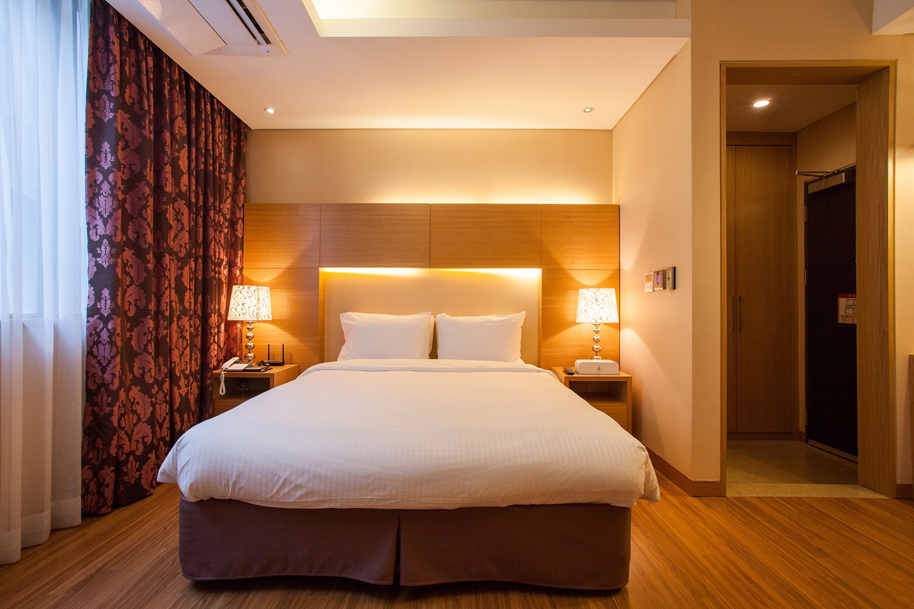 Une chambre d’hôtel avec un grand lit, deux tables de chevet avec des lampes, une tête de lit en bois et une porte donnant sur un couloir.