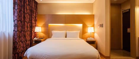 Une chambre d’hôtel avec un grand lit, deux tables de chevet avec des lampes, une tête de lit en bois et une porte donnant sur un couloir.