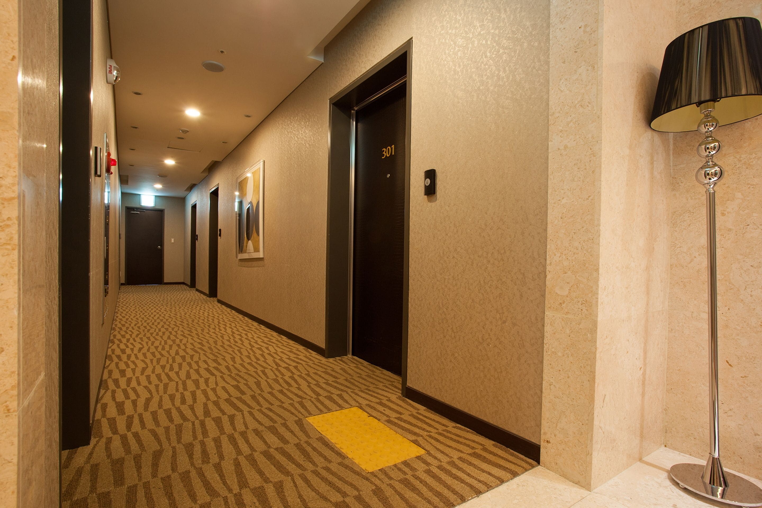 A hotel hallway with carpeted floors, doors, and a floor lamp.