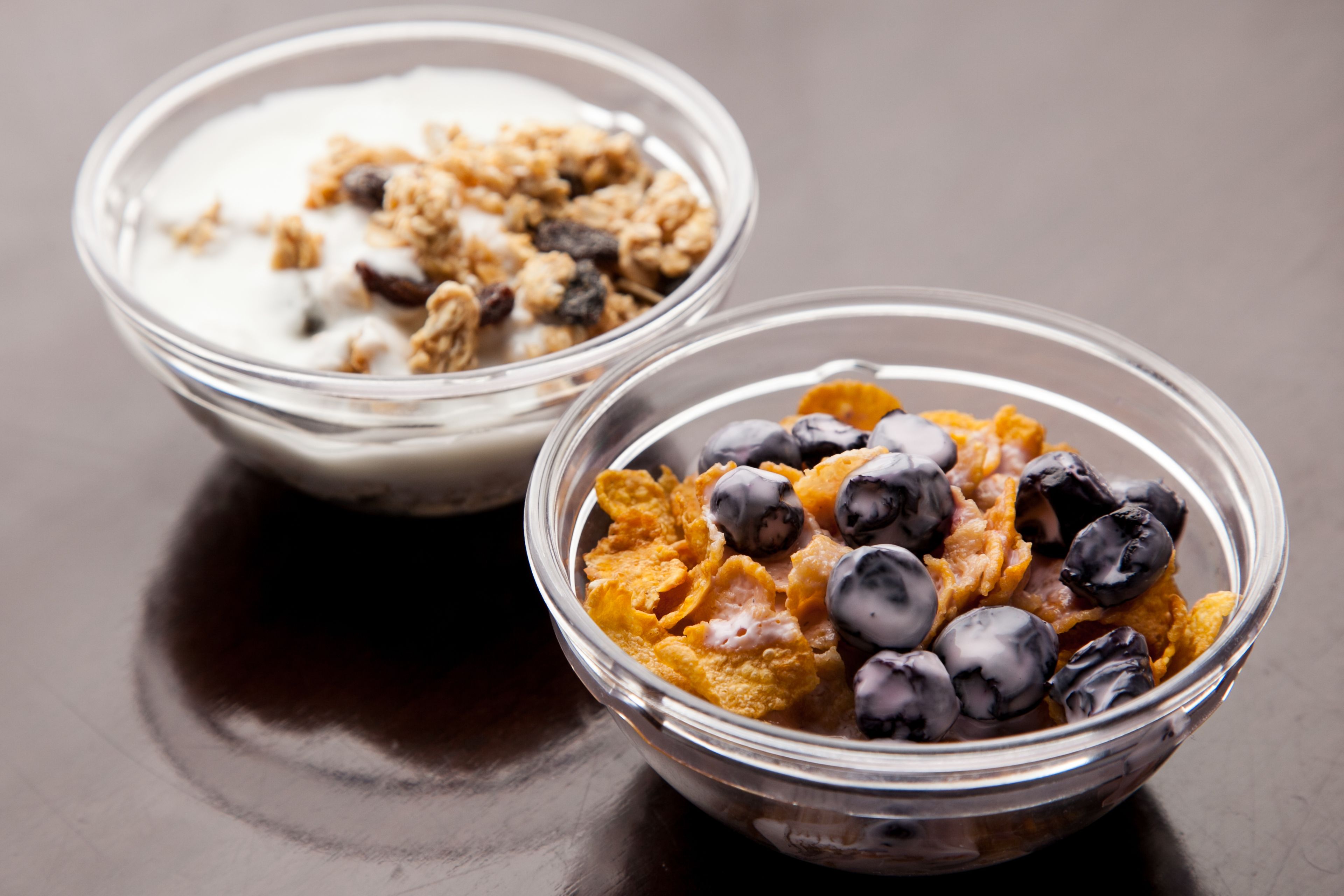 Dos tazones de vidrio transparente con distintos tipos de cereal, leche y fruta sobre una superficie oscura.