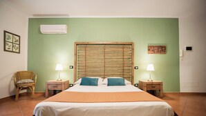 Una habitación con una cama grande, dos mesitas de noche con lámparas, una silla de mimbre, un aire acondicionado instalado en la pared y un pequeño cuadro en la pared.