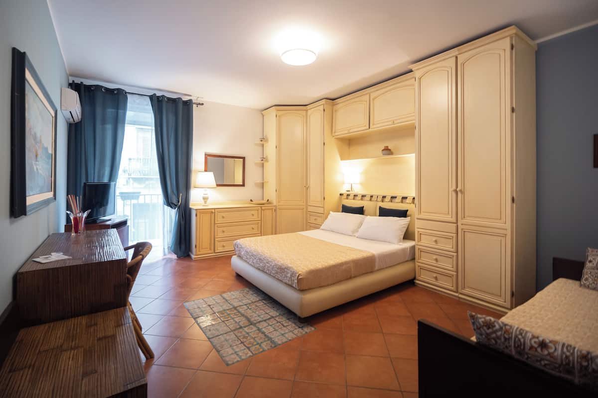 Une chambre à coucher avec un lit, un bureau et une chaise. Il y a une fenêtre avec des rideaux, une lampe et un placard.
