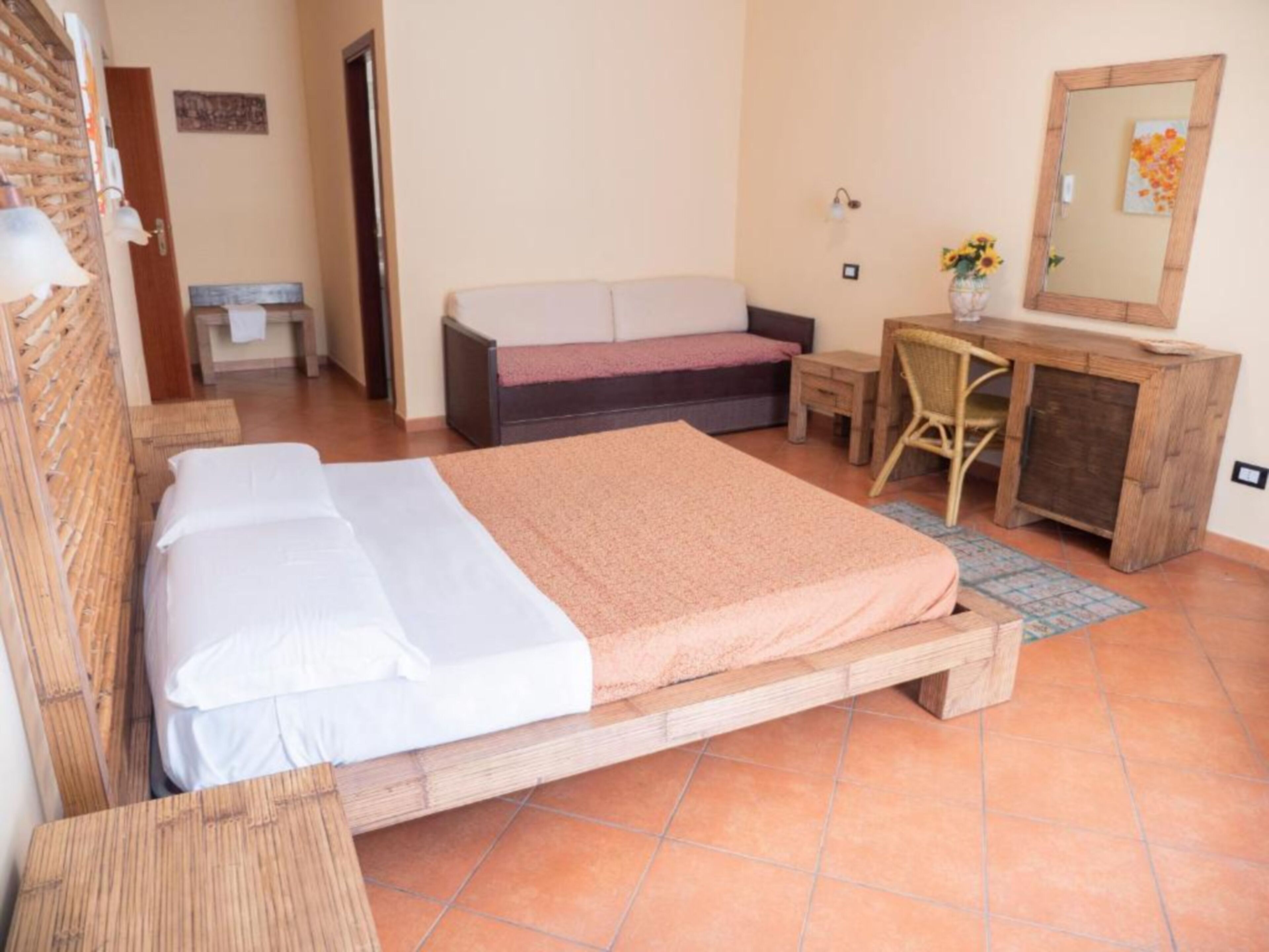 A bedroom with a bed, a small desk, a chair, a sofa, and a bathroom in the background.