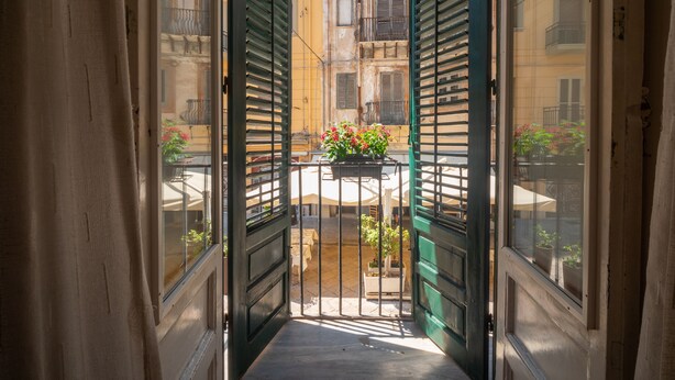 Un estrecho callejón con contraventanas verdes abiertas que da a un patio con mesas al aire libre y macetas con plantas.