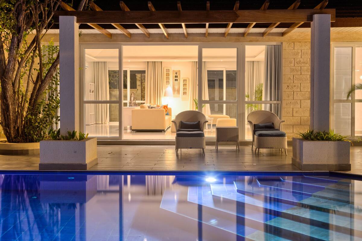 Zona junto a la piscina con un área de estar cubierta, piscina y vistas a un espacio interior bien iluminado con muebles y plantas.
