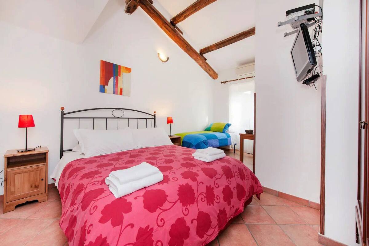 A bedroom with a large bed, a smaller bed, wooden nightstands, a wall-mounted TV, and a ceiling fan.