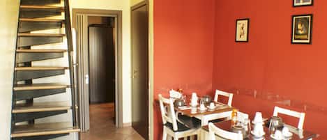 A dining area with a wooden table set for four, white chairs, and a red wall with framed pictures. A spiral staircase is visible to the left.