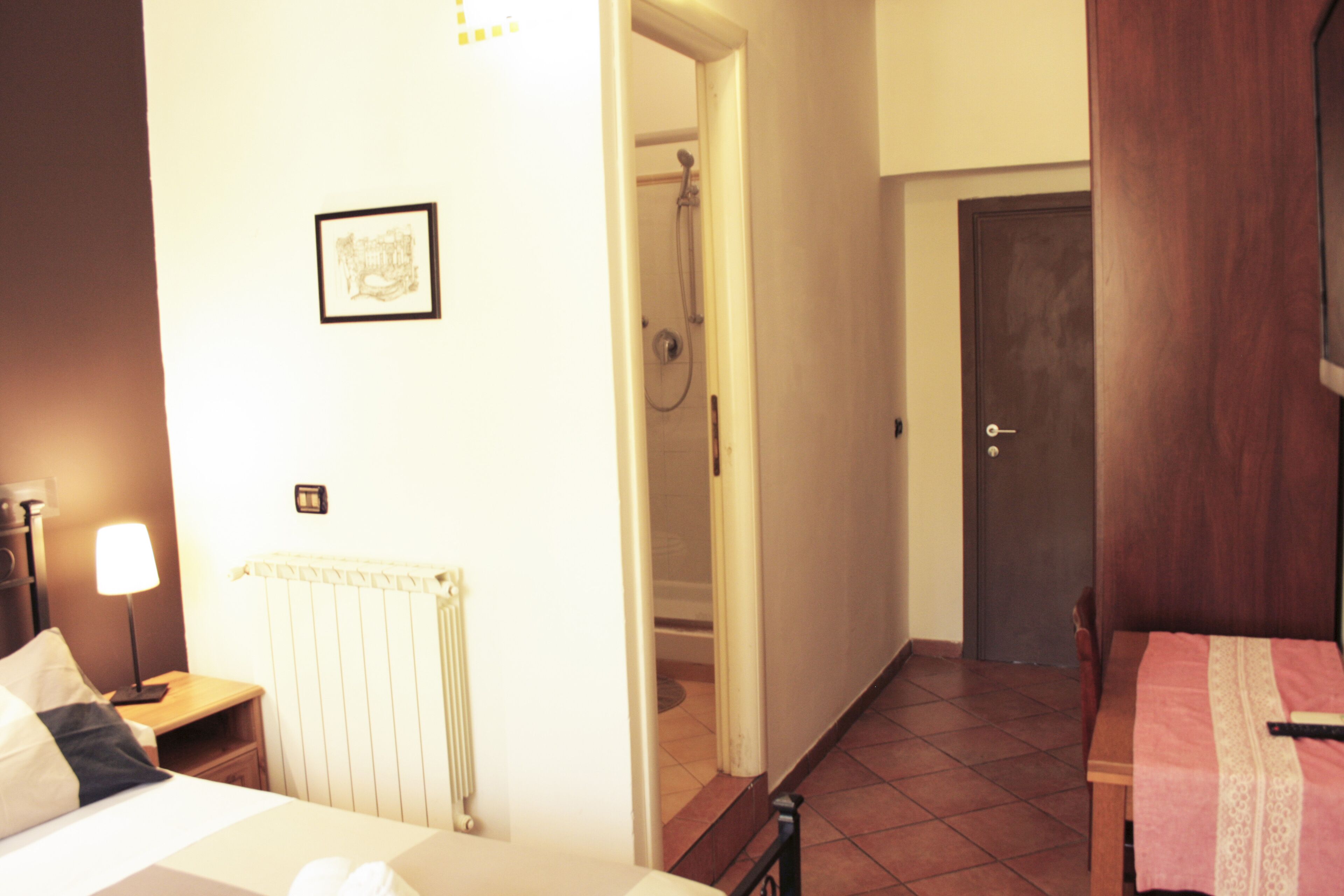 A hotel room with a bed, bedside table, lamp, radiator, shower, and a door.