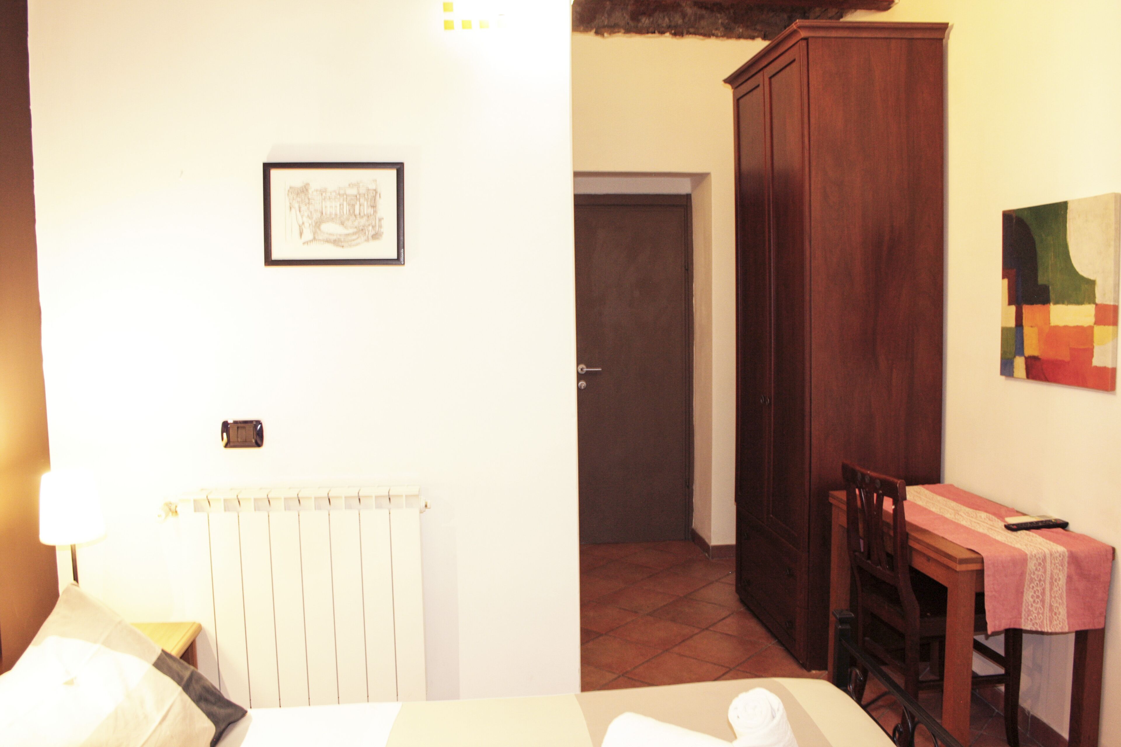A hotel room with a bed, a radiator, a wooden wardrobe, a dining table with a chair, and a framed picture on the wall.