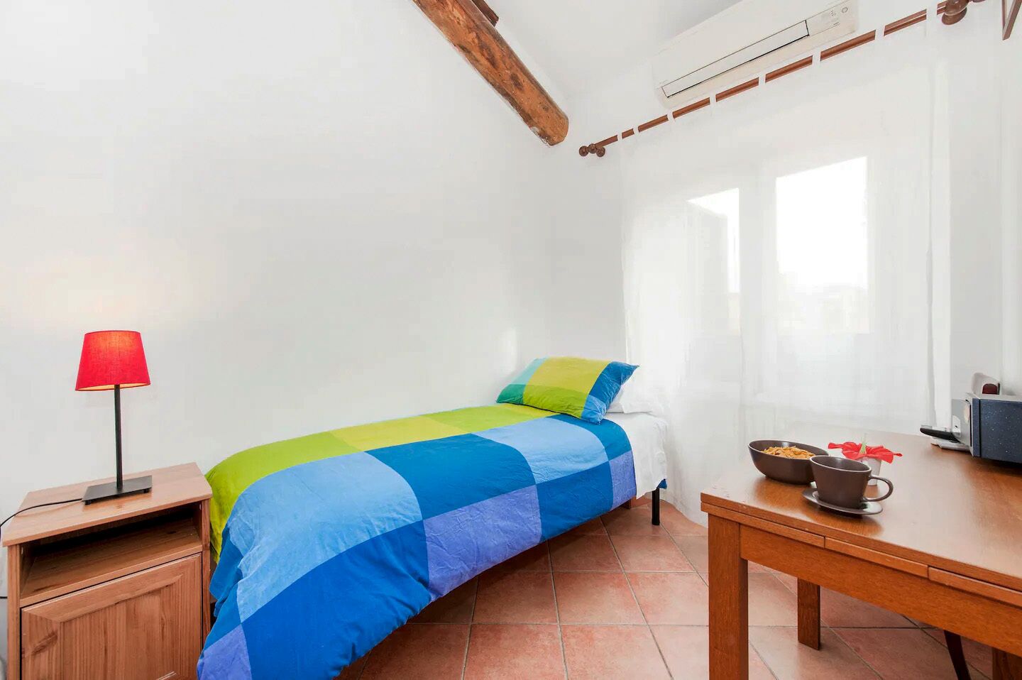 A bedroom with a bed, a nightstand, a table with a bowl and a cup, and a window with curtains.