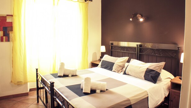 A neatly made bed with pillows and a folded towel, a bedside table with a lamp, and a window with curtains. - Sixtythree Guesthouse (Rome)