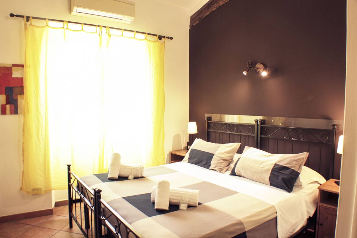 A neatly made bed with pillows and a folded towel, a bedside table with a lamp, and a window with curtains.