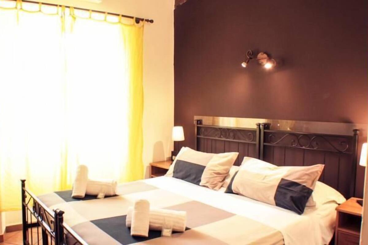 A hotel room with a bed, pillows, a nightstand, a lamp, and a window with curtains.