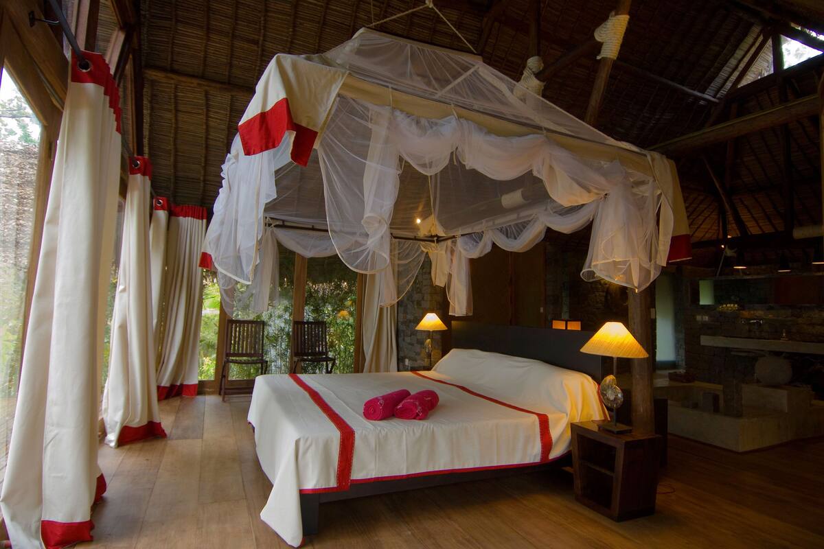 A room with a bed under a canopy, a nightstand with a lamp, and a stone wall.