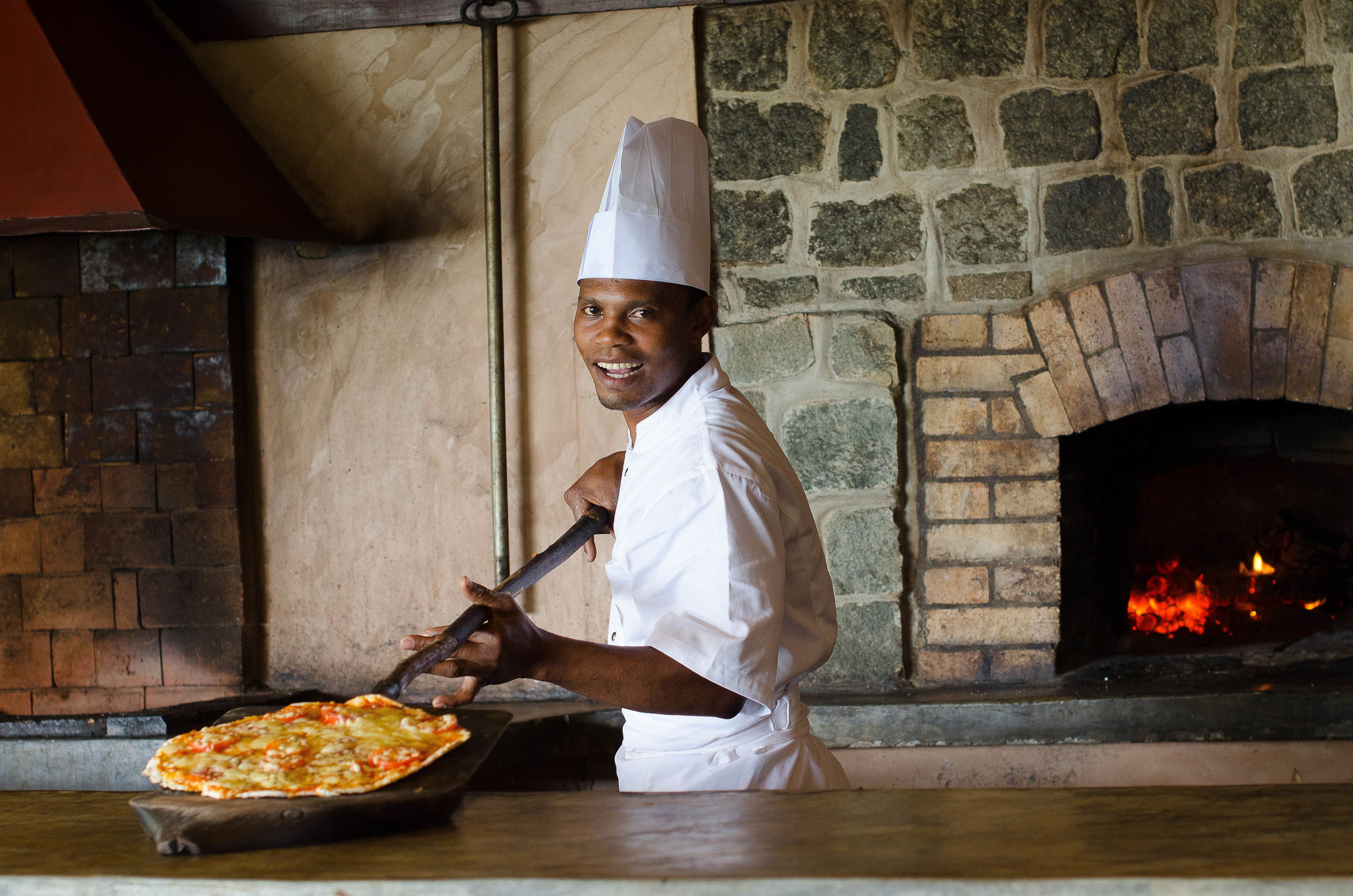 Un chef con uniforme y gorro blanco usa un utensilio de mango largo para introducir una pizza en un horno de ladrillo.