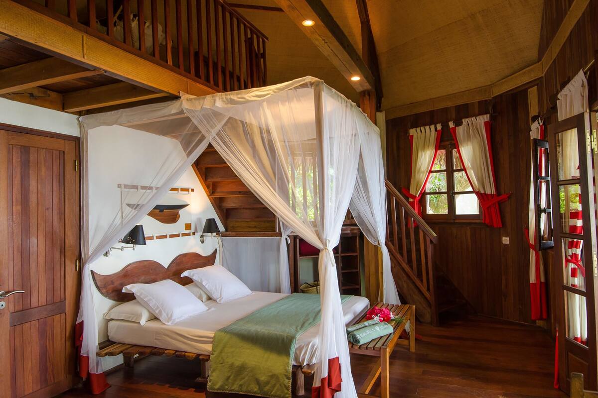 A wooden room with a bed under a canopy, a staircase, and a window with curtains.
