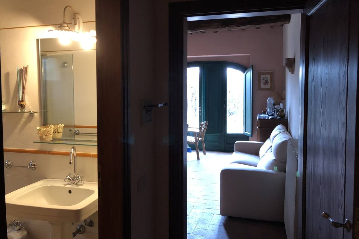 Ein Badezimmer mit Waschbecken, Spiegel und Handtuchhalter. Eine Tür führt zu einem Wohnzimmer mit Sofa und Tisch.