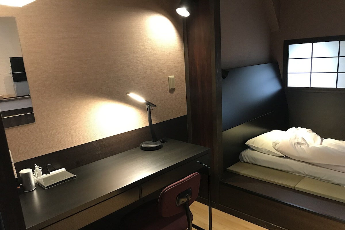 Ein kleines Hotelzimmer mit Schreibtisch, Stuhl, Nachttisch und einem Bett.