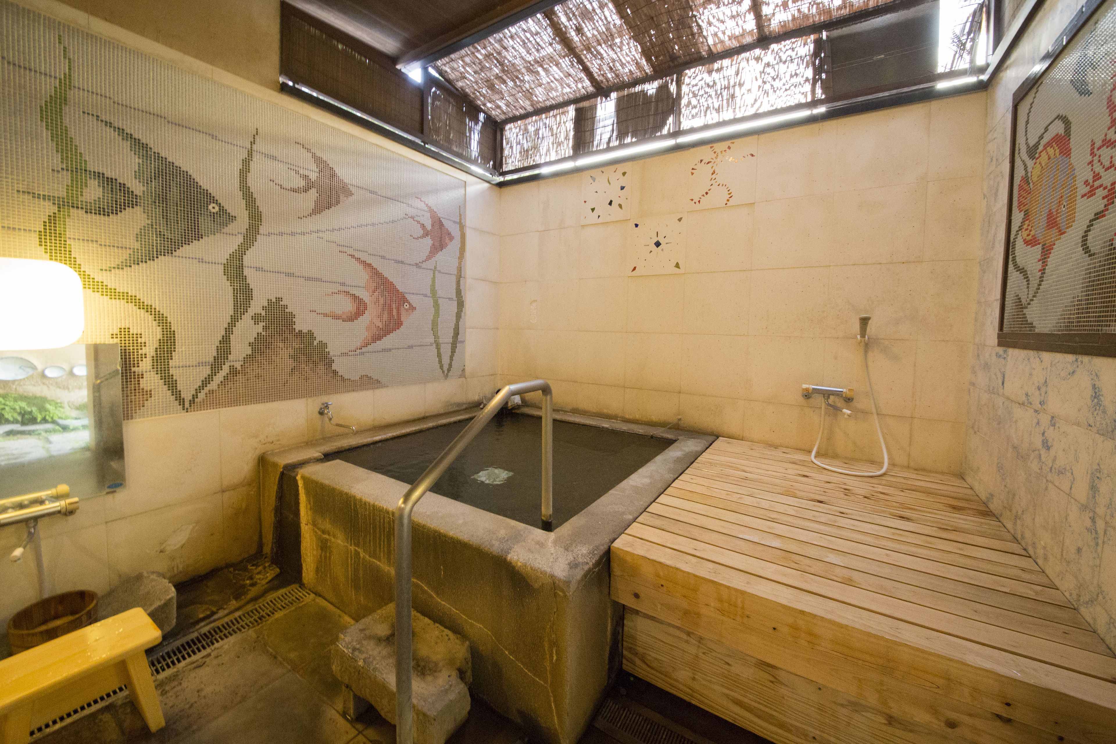A traditional Japanese onsen with a wooden bench, a stone hot spring, and tiled walls with fish and floral patterns.