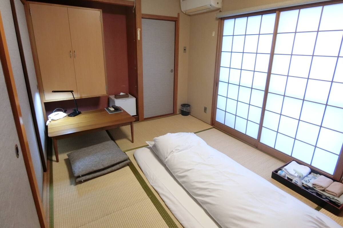 Ein traditioneller japanischer Raum mit Tatami-Bodenbelag, einer Futon-Liege, einem Holztisch mit Lampe und einer Schiebetür.