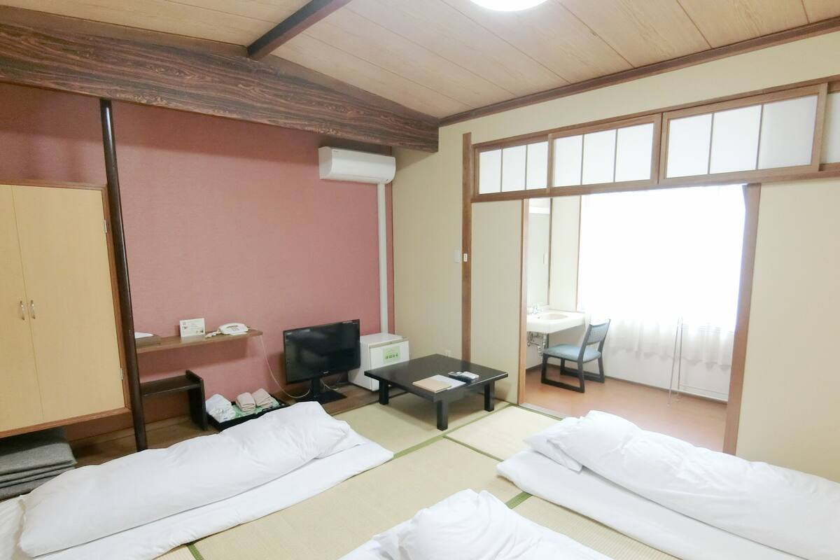 Ein traditioneller japanischer Raum mit Tatami-Böden, einem Fernseher, einem Schreibtisch und einem durch eine Schiebetür einsehbarer Badezimmer.