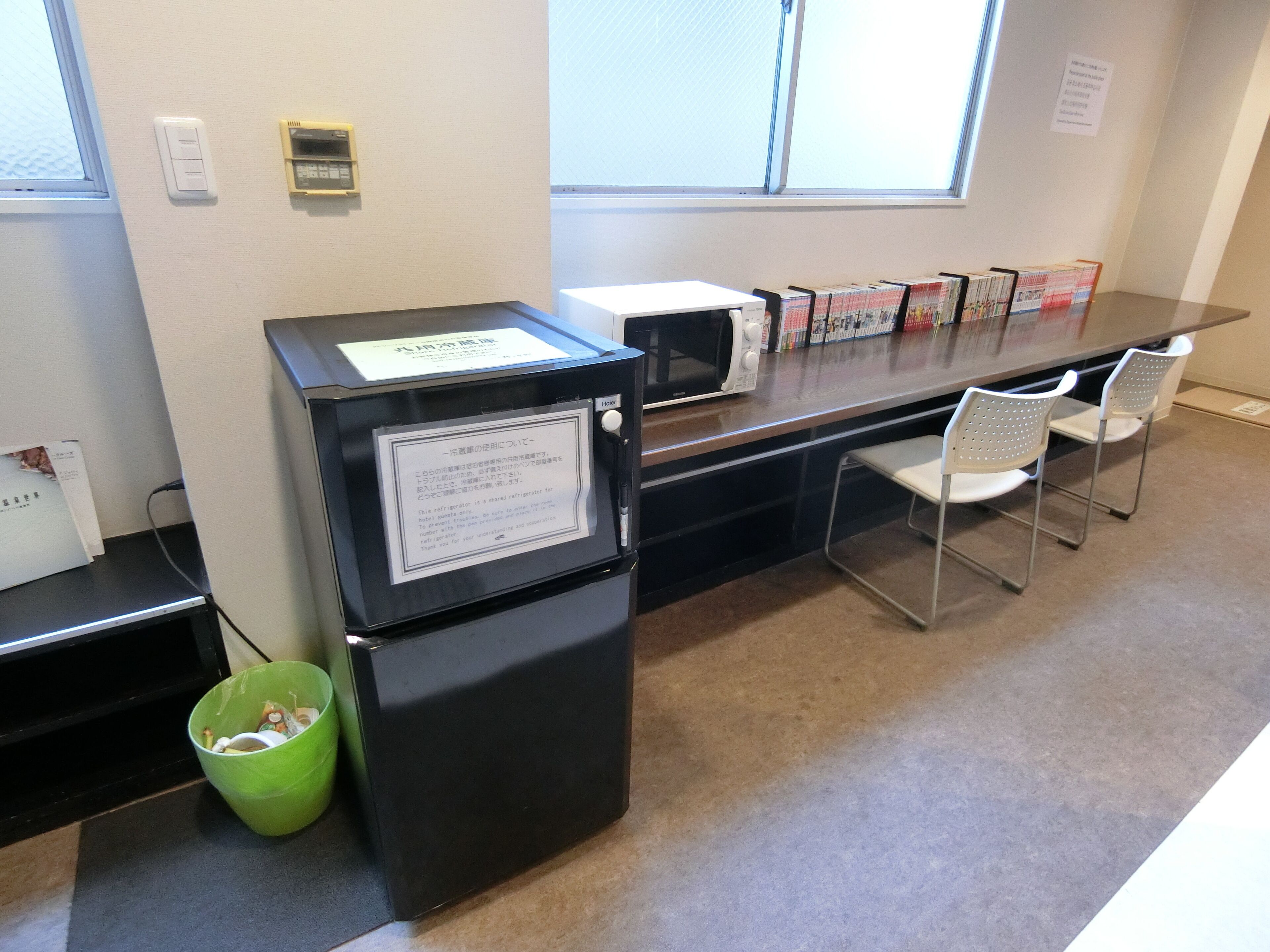 A hotel room with a kitchenette, a microwave, a refrigerator, and a trash bin. There are two chairs and a table with magazines.