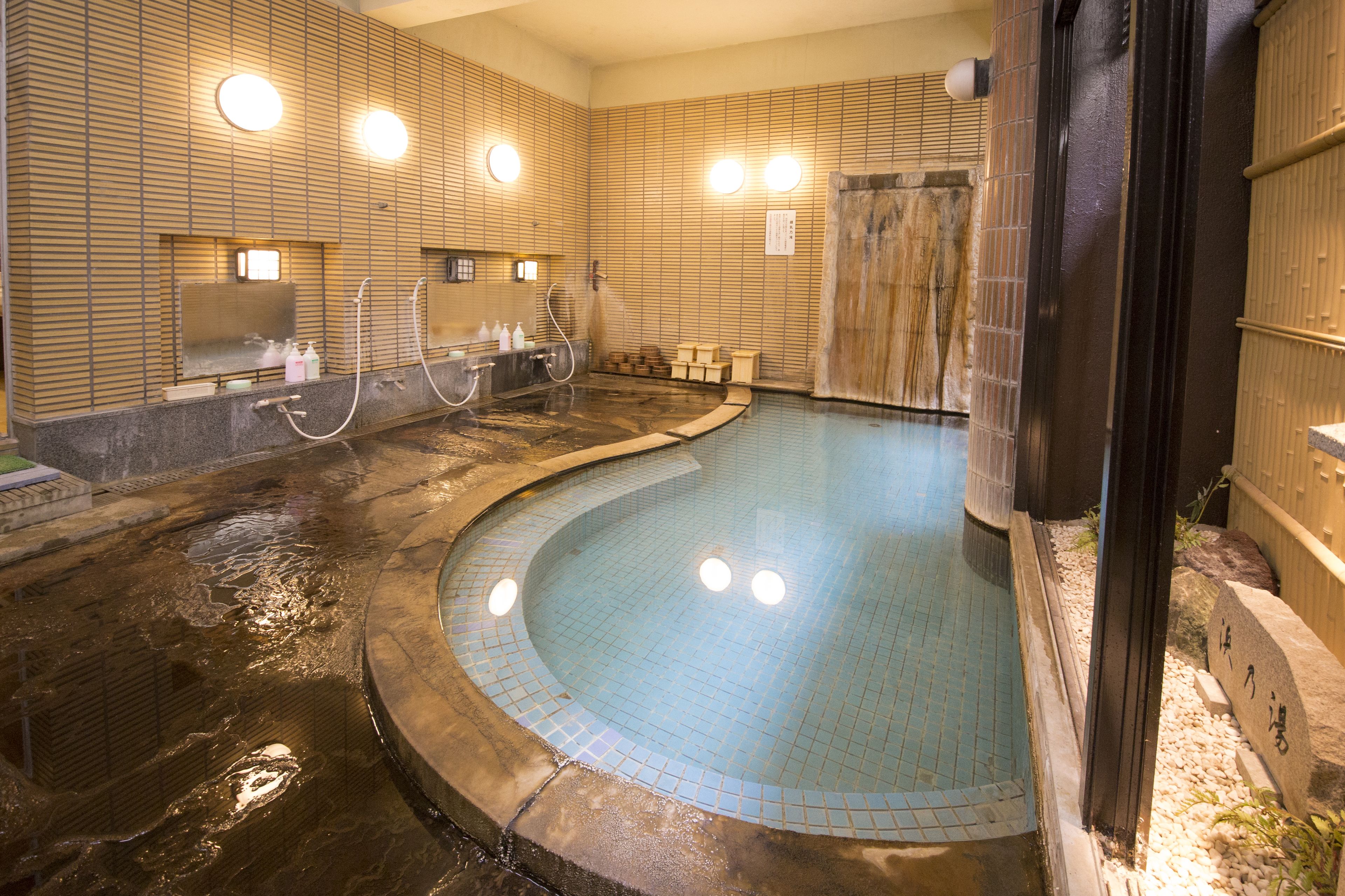 Un área de baño de aguas termales con paredes de azulejos, múltiples grifos y una piscina curva.