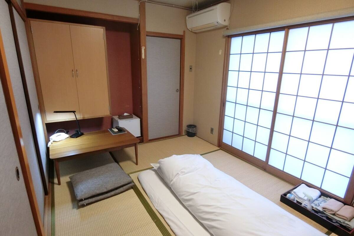 Ein traditioneller japanischer Raum mit Tatami-Bodenbelag, einem Futonbett, einem Holztisch mit Lampe, einem Kleiderschrank und einer schiebbaren Shoji-Schiebetür.
