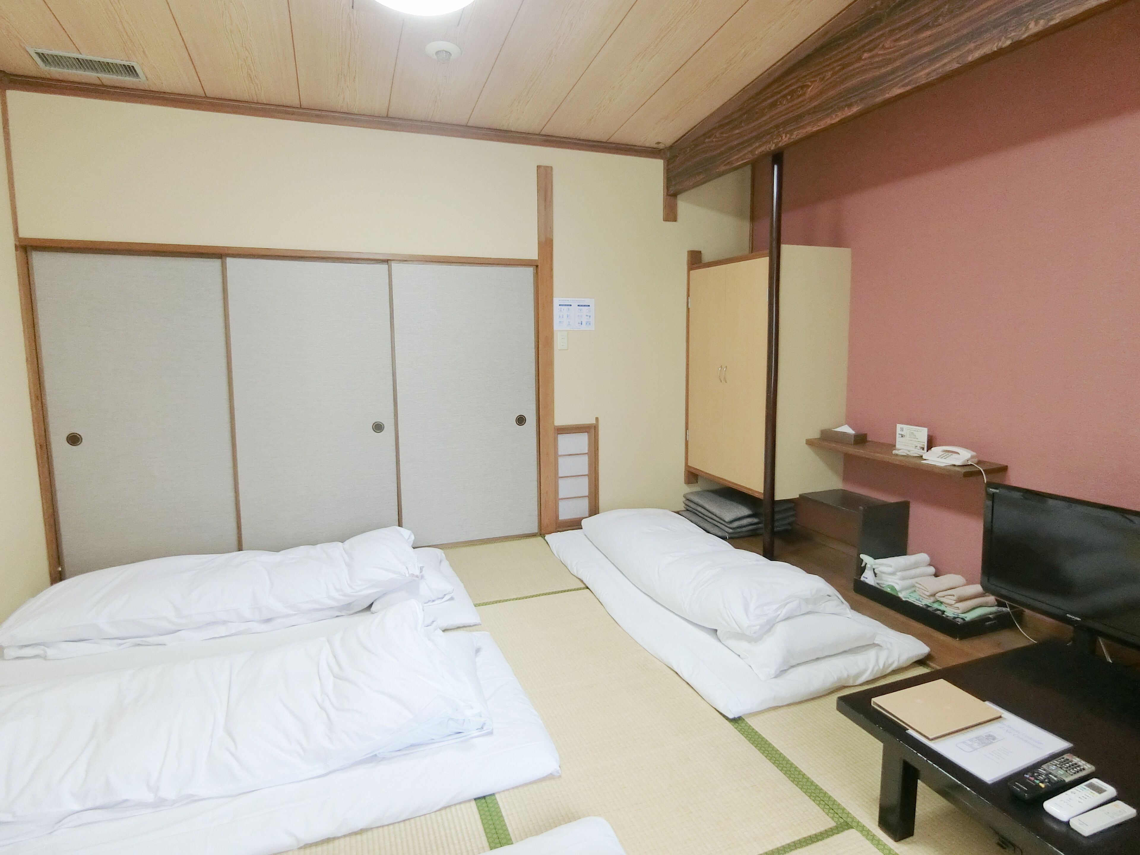 Une chambre d’hôtel avec des tatamis, un lit avec du linge de lit blanc, un bureau avec une télévision et une étagère avec des serviettes.