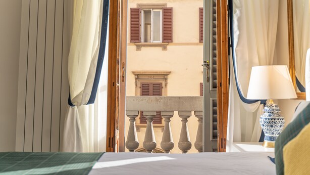 A hotel room with a balcony, a bed, a nightstand with a lamp, and a view of a building with shutters. - La Residenza di Michelangelo (Florence)