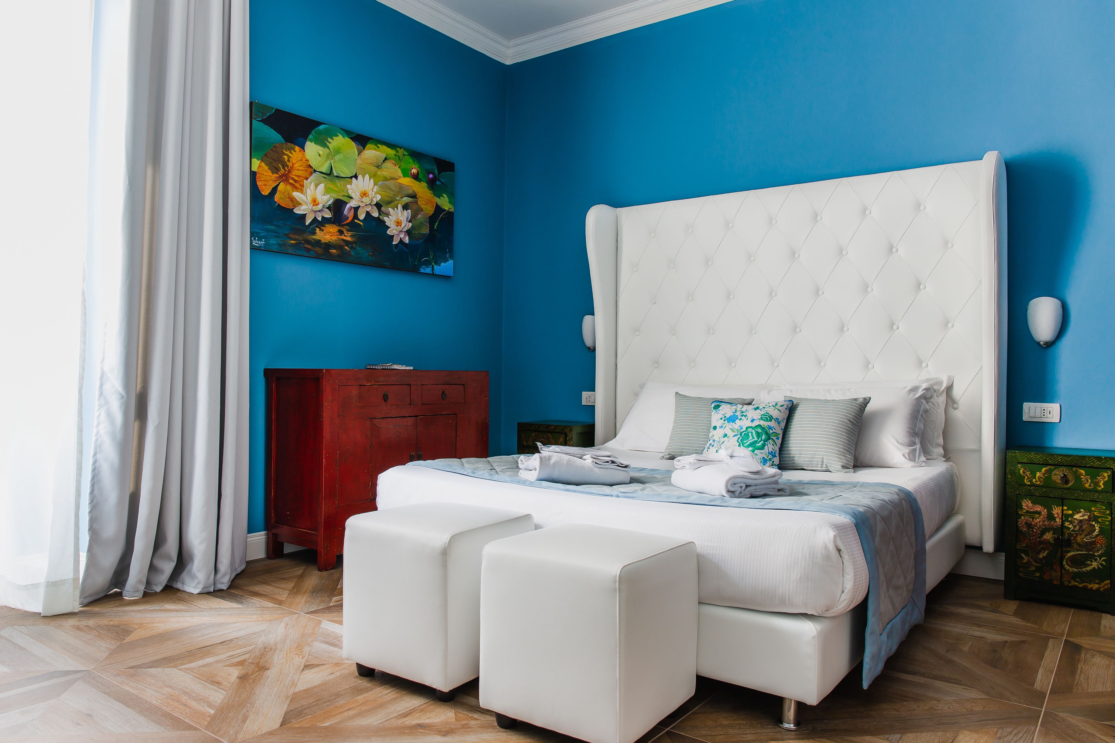 A bedroom with a large bed, two white ottomans, a red cabinet, and a painting of flowers on the wall.