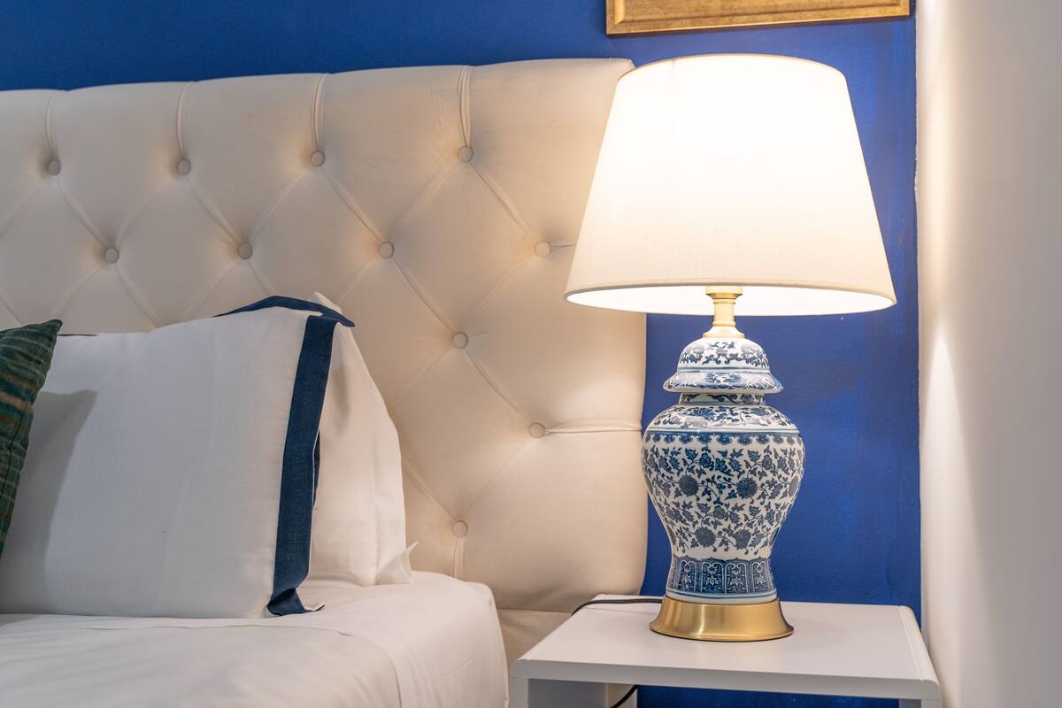 A neatly made bed with a white headboard, a blue and white decorative lamp on a white nightstand, and a blue wall in the background.