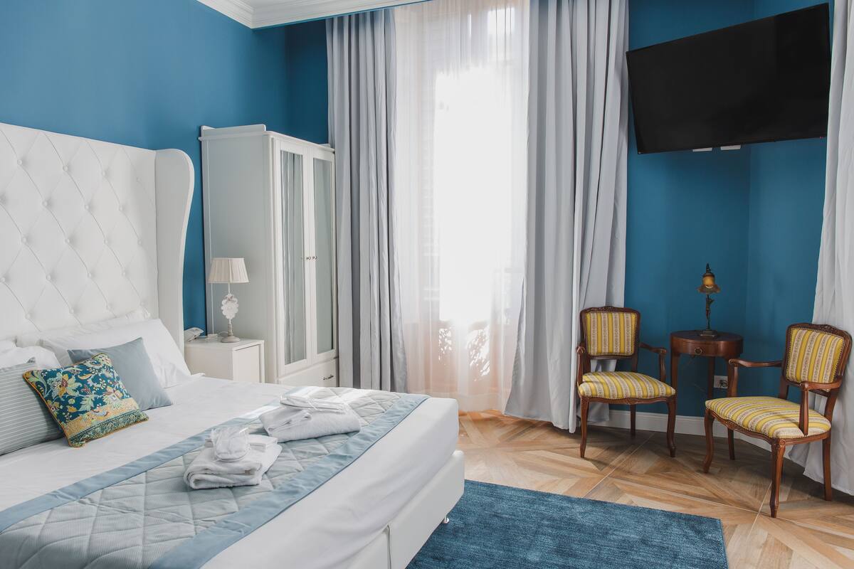 A bedroom with a bed, a wardrobe, two chairs, a TV, and a window with curtains.