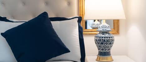 A bed with a tufted headboard, a navy blue pillow, a white pillow, and a bedside table with a blue and white ceramic lamp.