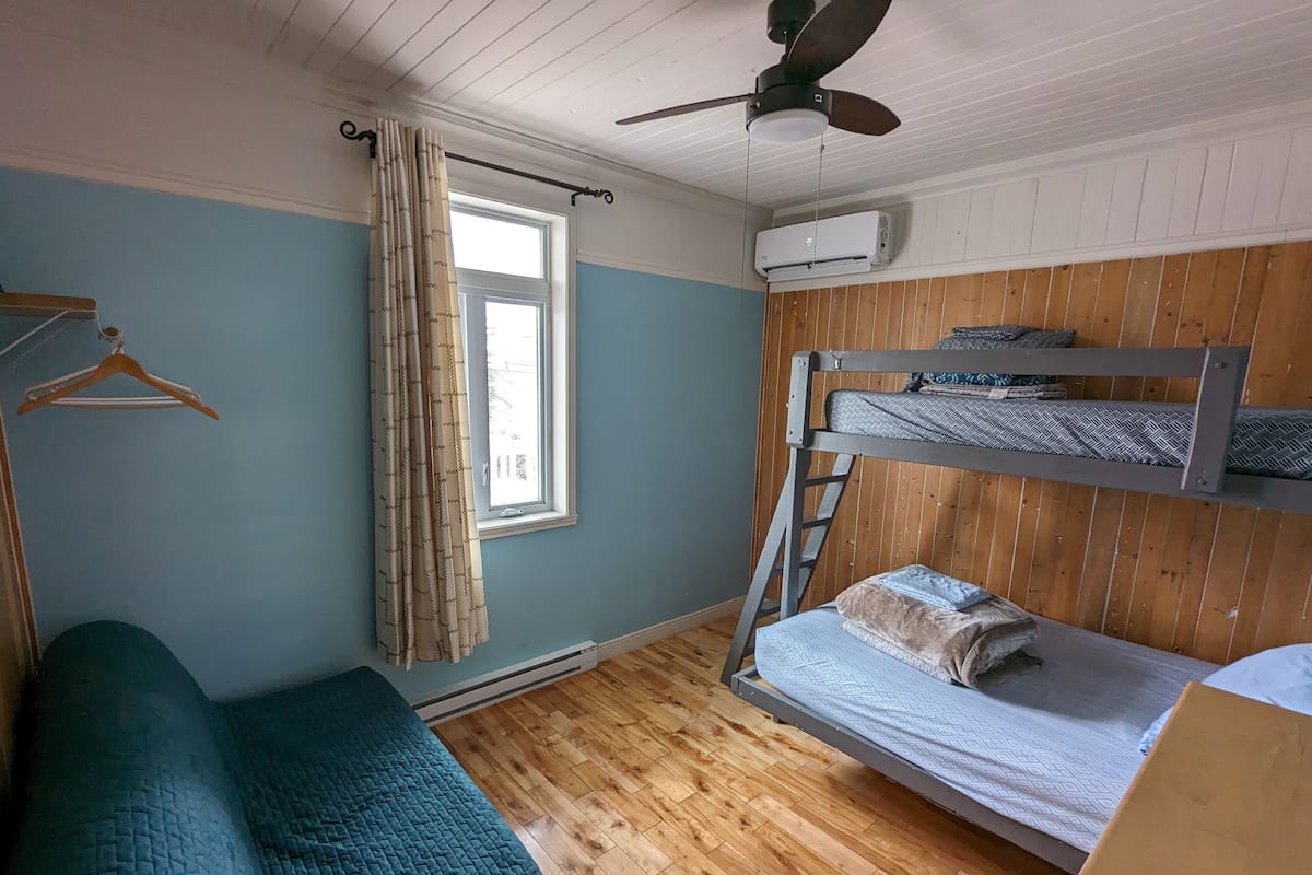 Una stanza con un letto a castello, un letto singolo, una ventola a soffitto, una finestra con tende e un condizionatore d'aria.