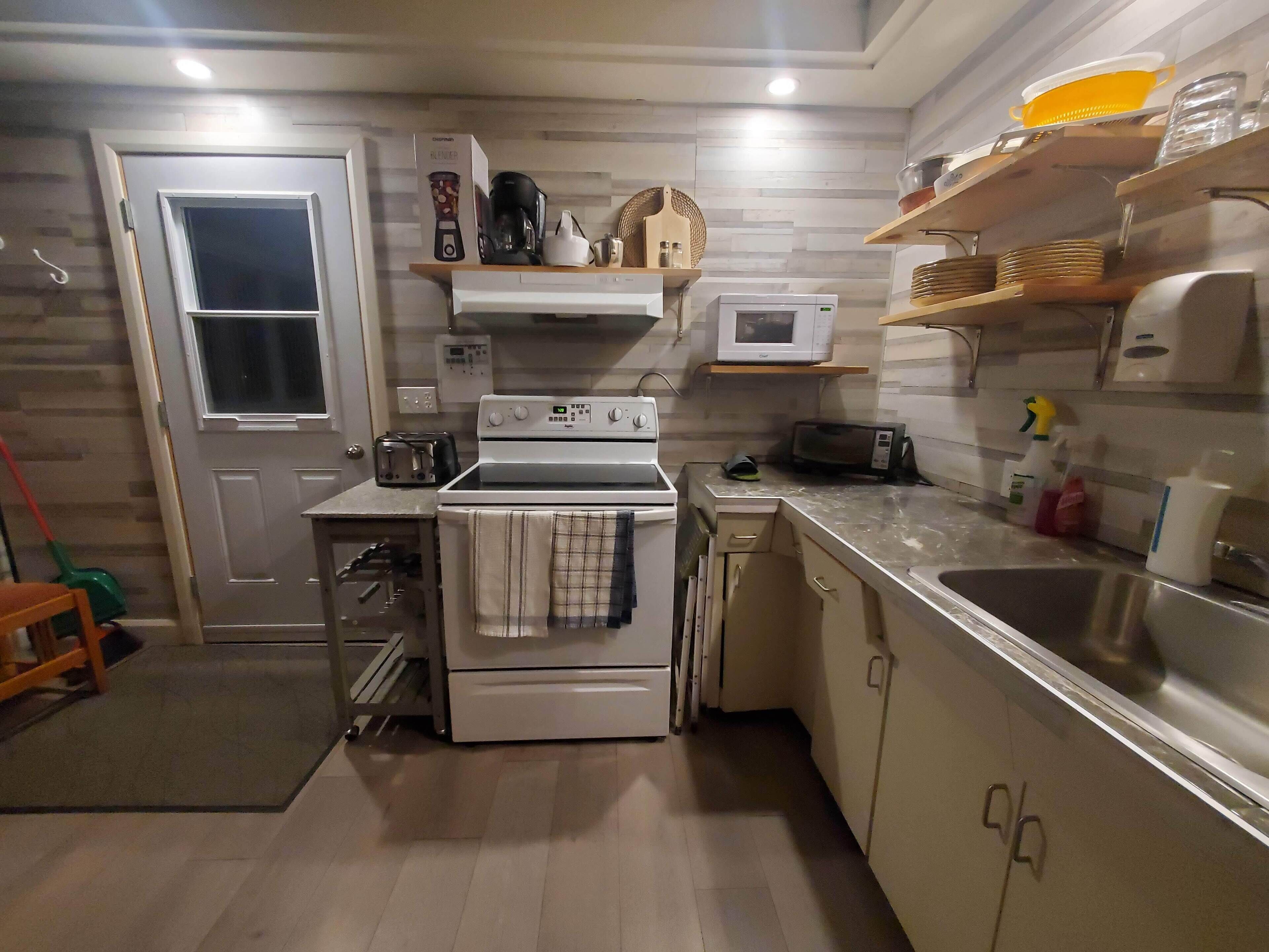 Una cocina compacta con estufa, microondas y tostadora. Hay repisas de madera sobre la encimera con vajilla y una cesta. Se ve una puerta con ventana a la izquierda.